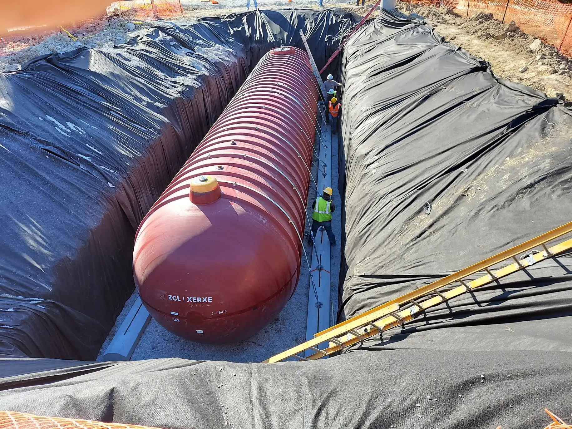 A large red fiberglass underground storage tank set in a lined excavation, with crew working alongside it