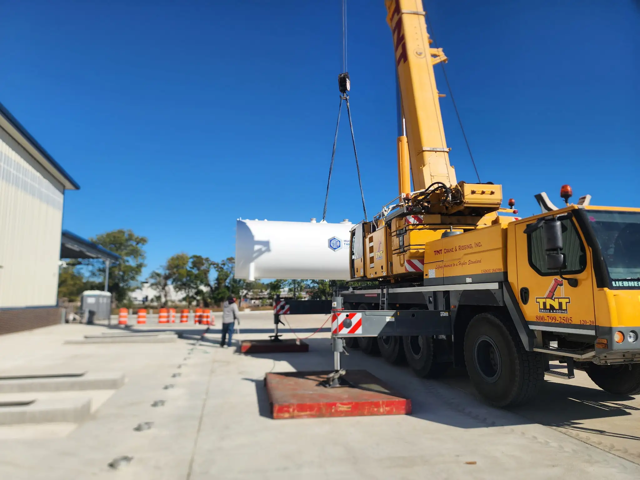 A crane lowers a tank toward its foundation pad as a crew member guides it into position